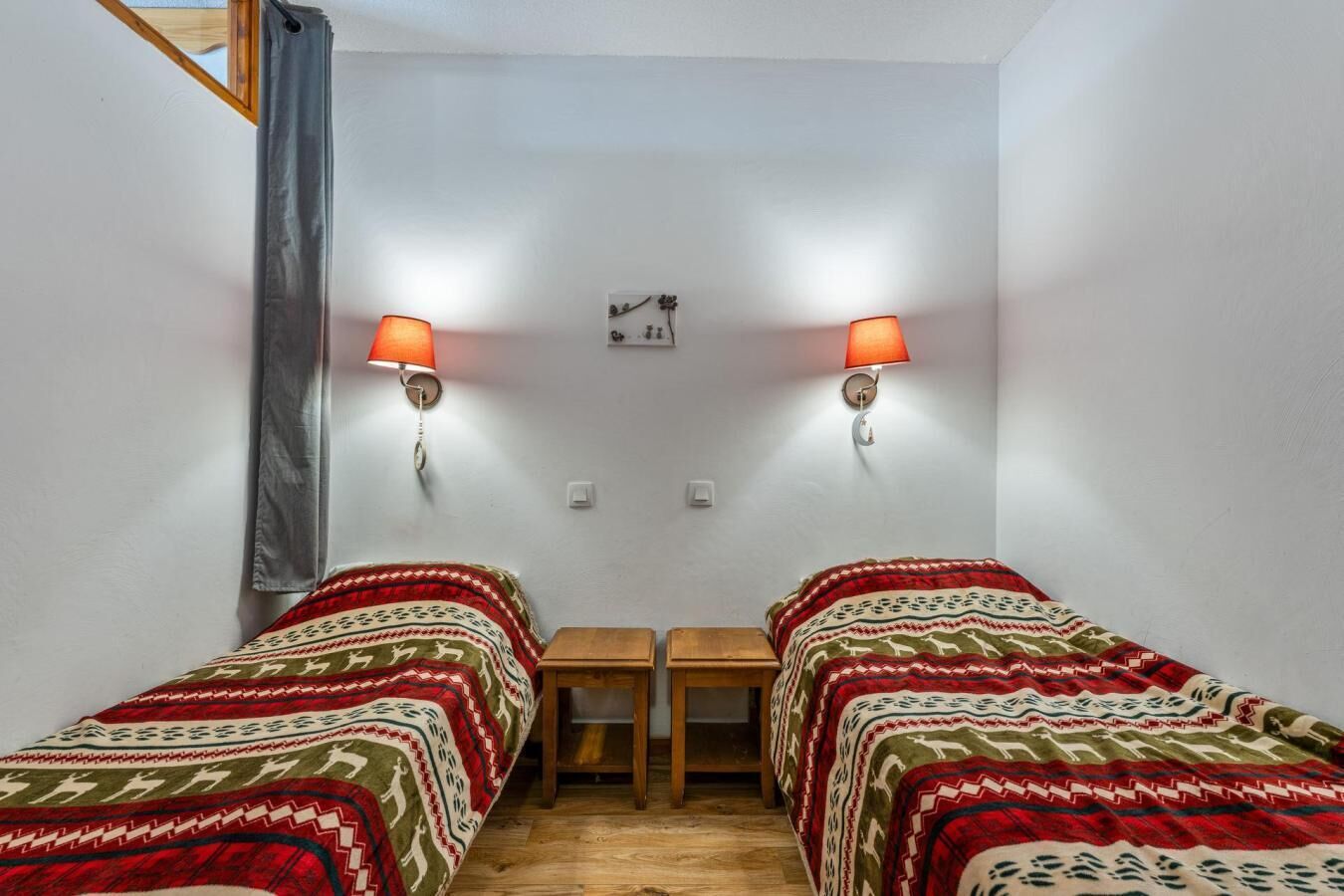Quarto pequeno e simples com duas camas de solteiro, uma mesinha e duas luminárias.