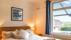 Una cama bien hecha con sábanas y almohadas blancas, una lámpara de noche y una ventana con vistas a zonas verdes y un edificio.