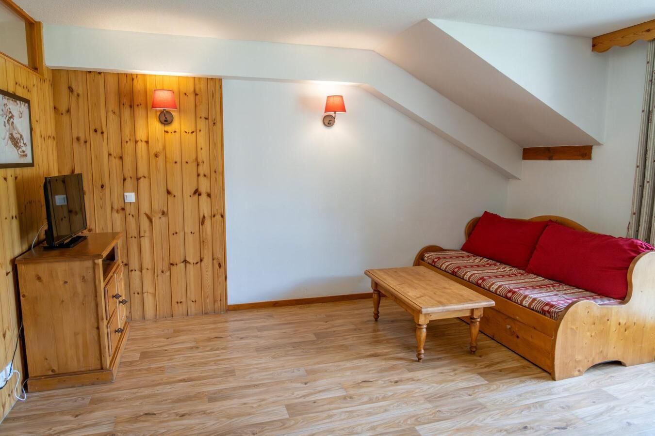 Una habitación acogedora con pared de madera, un sofá con cojines rojos, una mesa de madera y un televisor sobre un mueble.
