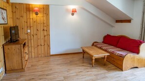 A cozy room with a wooden wall, a sofa with red cushions, a wooden table, and a TV on a cabinet. - 40m from ski slopes, 3rd floor, fitness, swimming pool, sauna, balcony, tv, ski locker, 30m² (Les Orres)