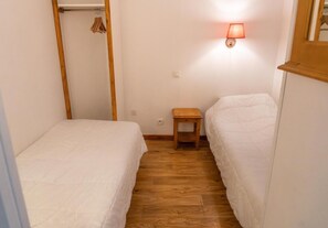 A small, narrow room with two single beds, a wooden bench, and a wall-mounted light fixture.