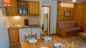 Une cuisine compacte avec des meubles en bois, une machine à café et un lave-vaisselle. On y trouve une table à manger avec des chaises, ainsi qu’un canapé dans l’espace salon.