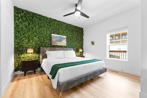 Quarto com cama, mesas de cabeceira, parede verde, ventilador de teto e janela com vista para varanda.