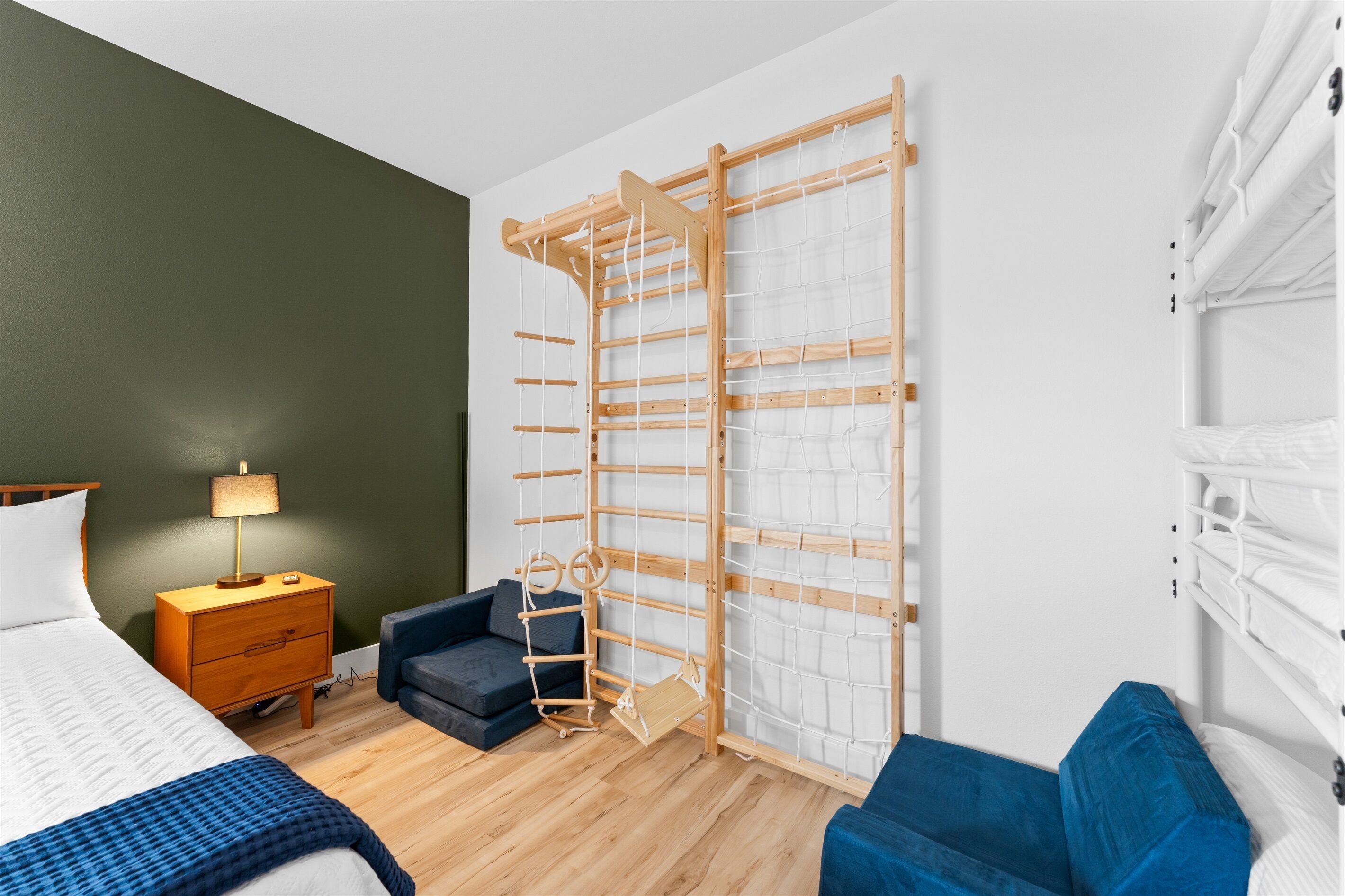 A bedroom with a bed, a nightstand, a blue sofa, a wooden climbing wall, and a white bunk bed.