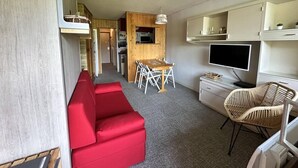 Un espacio habitable compacto con un sofá rojo, una mesa de comedor con sillas, un televisor y una pequeña cocina.