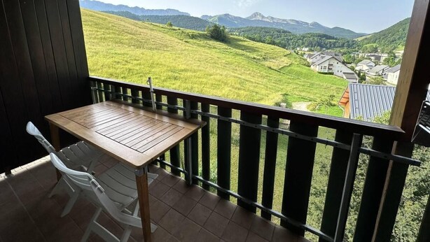 Un balcón con una mesa de madera y sillas blancas, con vista a un valle verde con casas y montañas.