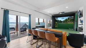 A modern room with a large screen displaying a golf game, a balcony with a view, and a dining area with chairs. - Branson GOAT: Golf Simulator Hot Tub Lake View Fun (Branson)