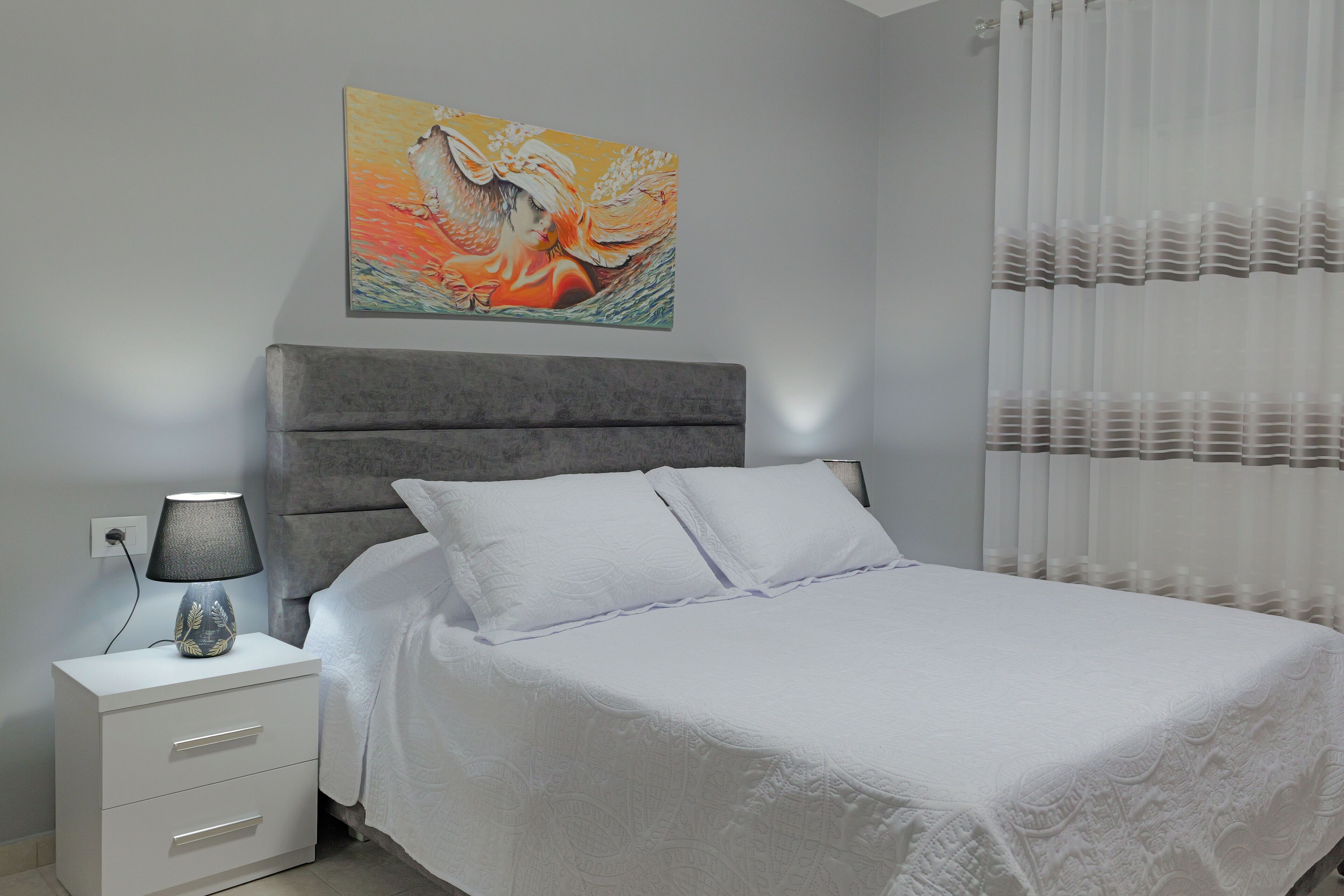 A bedroom with a bed, a nightstand, a lamp, a painting, and a window with curtains.