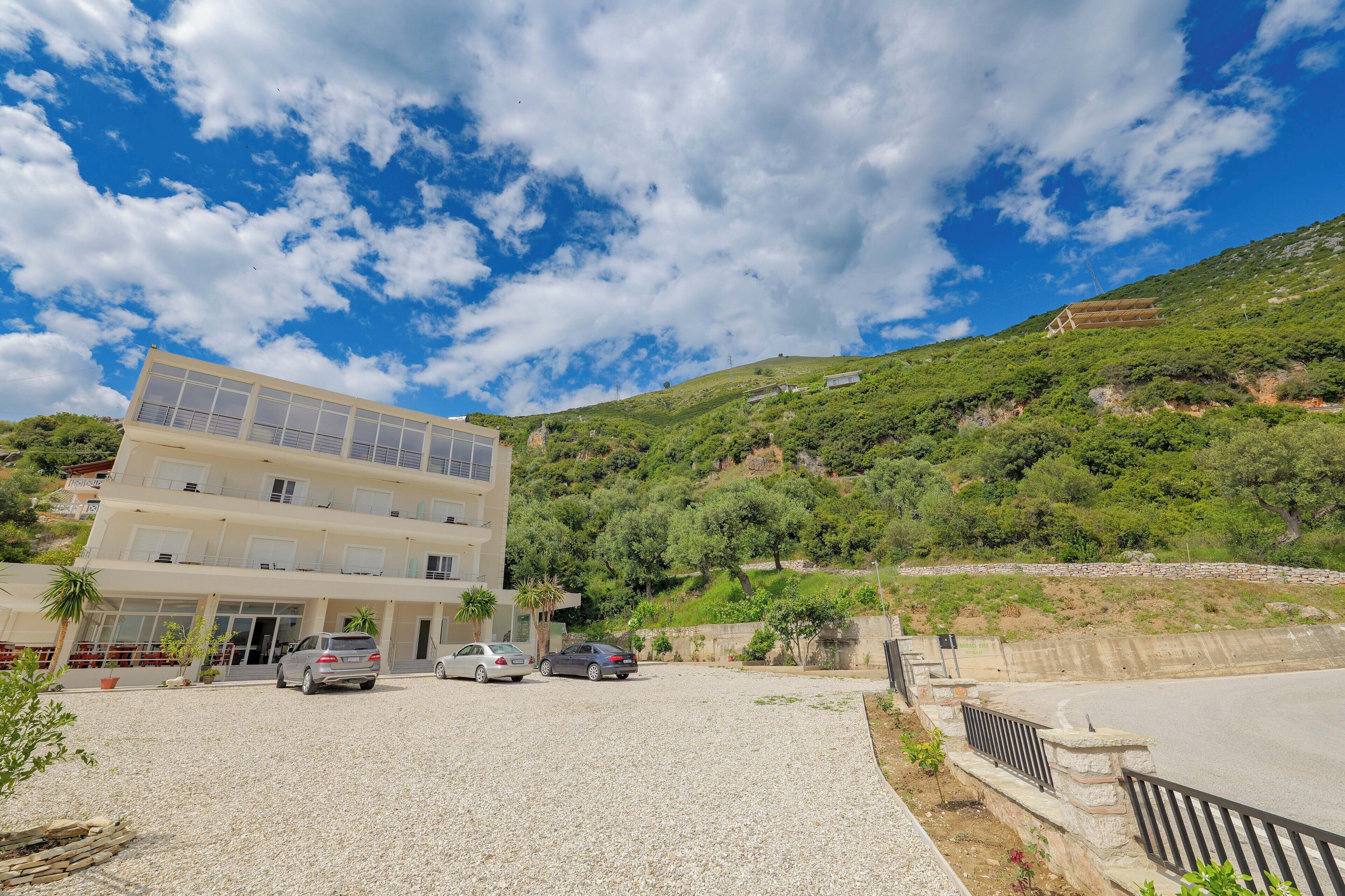 A multi-story hotel with a parking area, a hilly backdrop, and a clear sky.