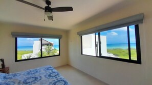 A bedroom with a bed, a ceiling fan, and two large windows offering views of the ocean and a building.