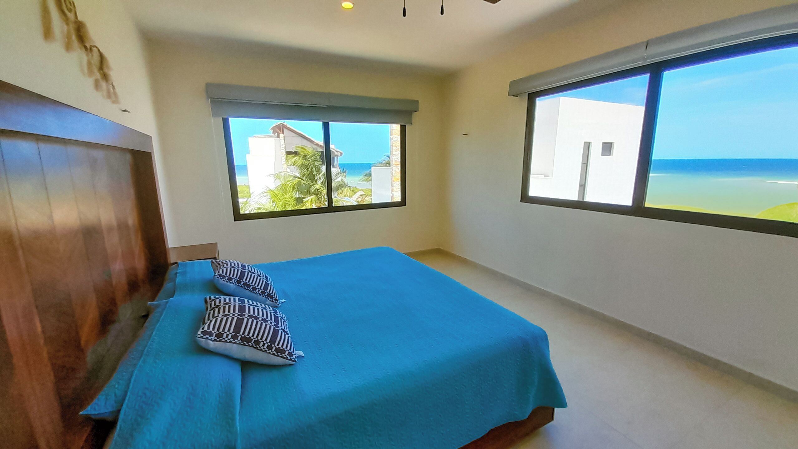 A bedroom with a bed, two pillows, a window with a view of the beach, and a ceiling fan.