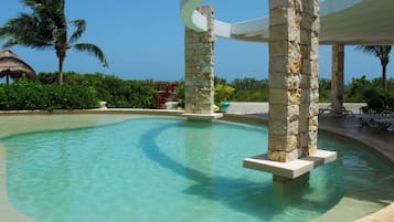 Una piscina cristalina con pilares de piedra, una palmera y una estructura de techo de paja.