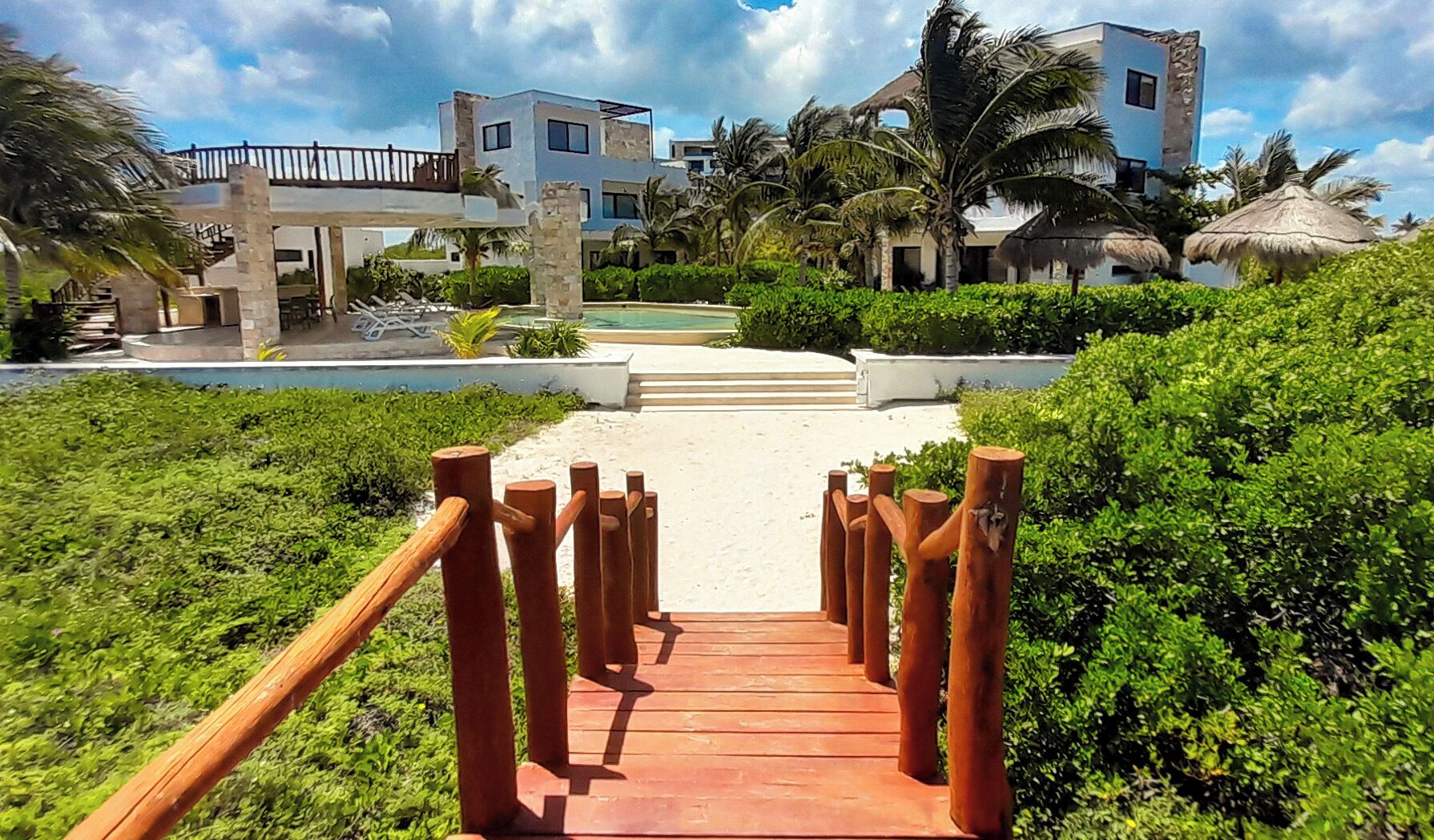 Una escalera de madera que conduce a una zona de playa con piscina y edificios modernos al fondo.