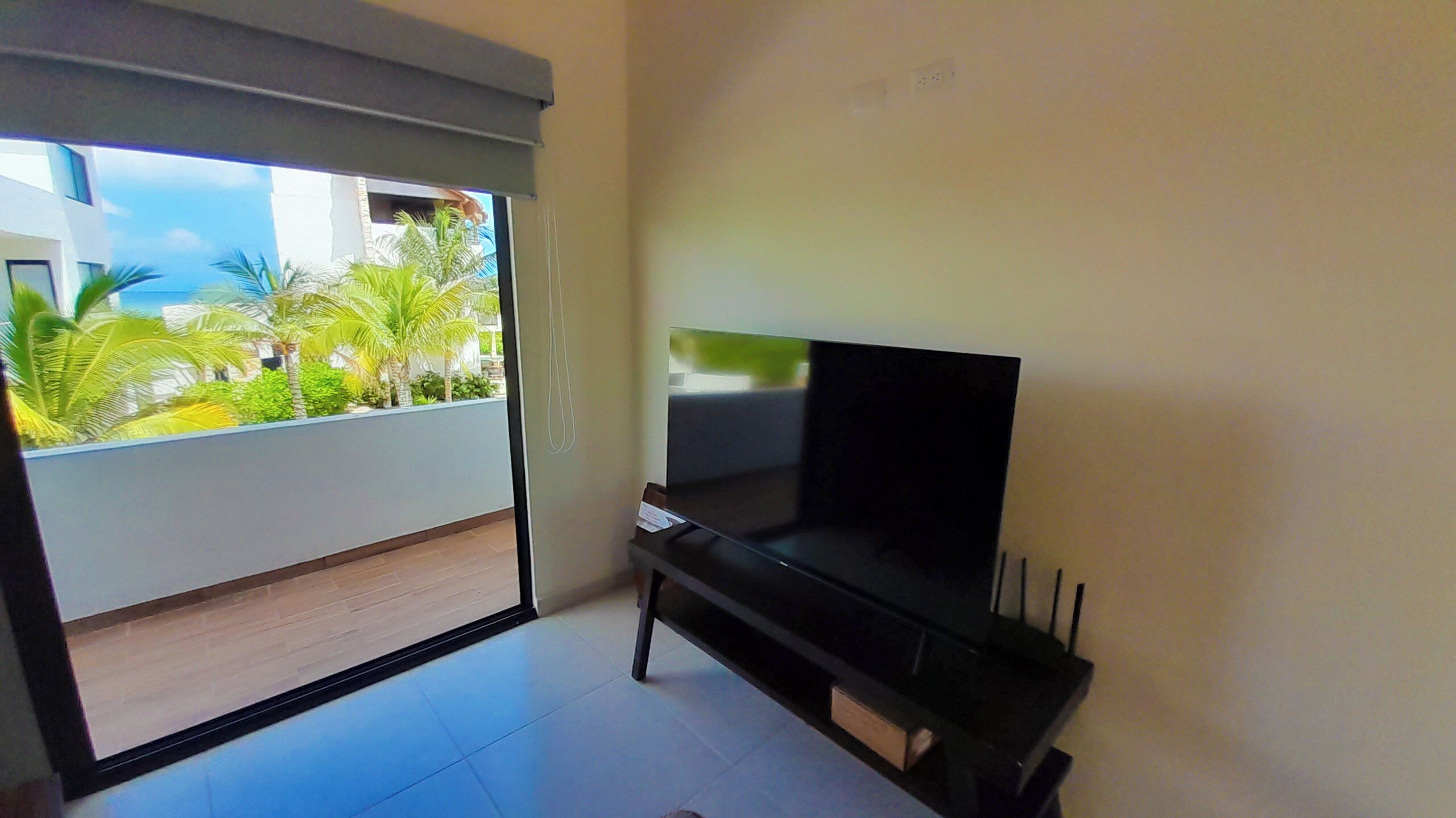 Una sala de estar con televisor, puerta corrediza de vidrio a un balcón con plantas y piso de baldosas.