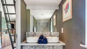 Een badkamer met twee wasbakken, een marmeren aanrecht, een handdoekrek met opgerolde handdoeken, een spiegel en een kunstwerk in een lijst aan de muur.