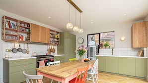 Une cuisine avec une table en bois, des chaises et un fourneau. On y trouve des placards, une horloge et une fenêtre donnant sur l’extérieur.