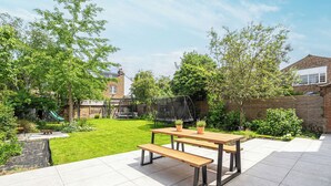 Une terrasse avec une table et des bancs en bois, entourée d’une pelouse bien entretenue et d’arbres.