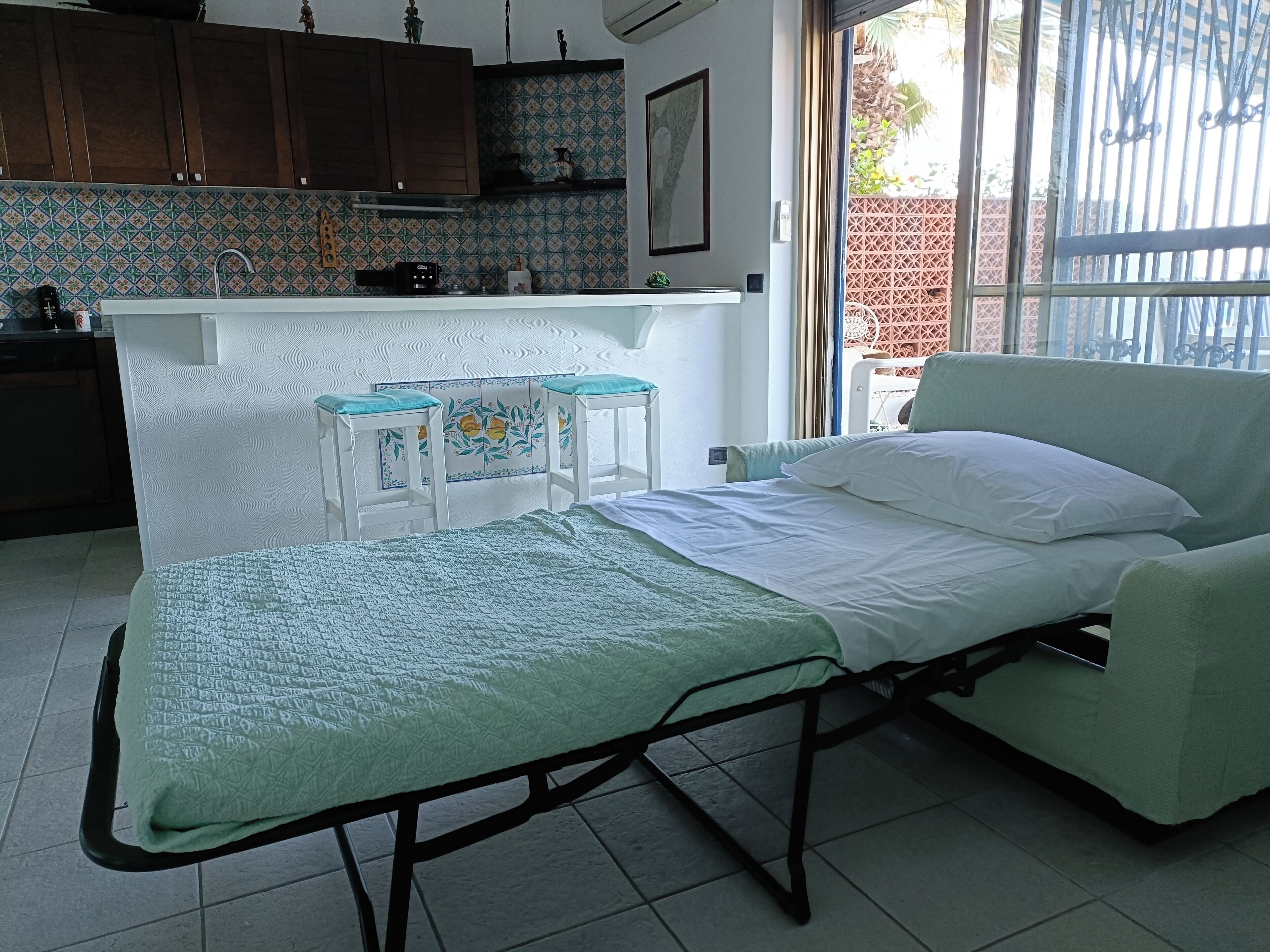 A single-bed room with a green bedspread, a bedside table, and a chair. There is a kitchenette with a sink, a stove, and a small refrigerator. The room has a tiled floor and a window with a view of a fence and greenery outside.