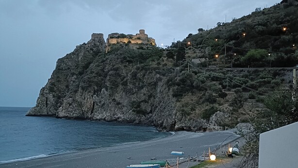 A coastal road with boats, a rocky cliff, and a castle-like structure in the background. - Margarita Ocean View Apartment (Sant'Alessio Siculo)