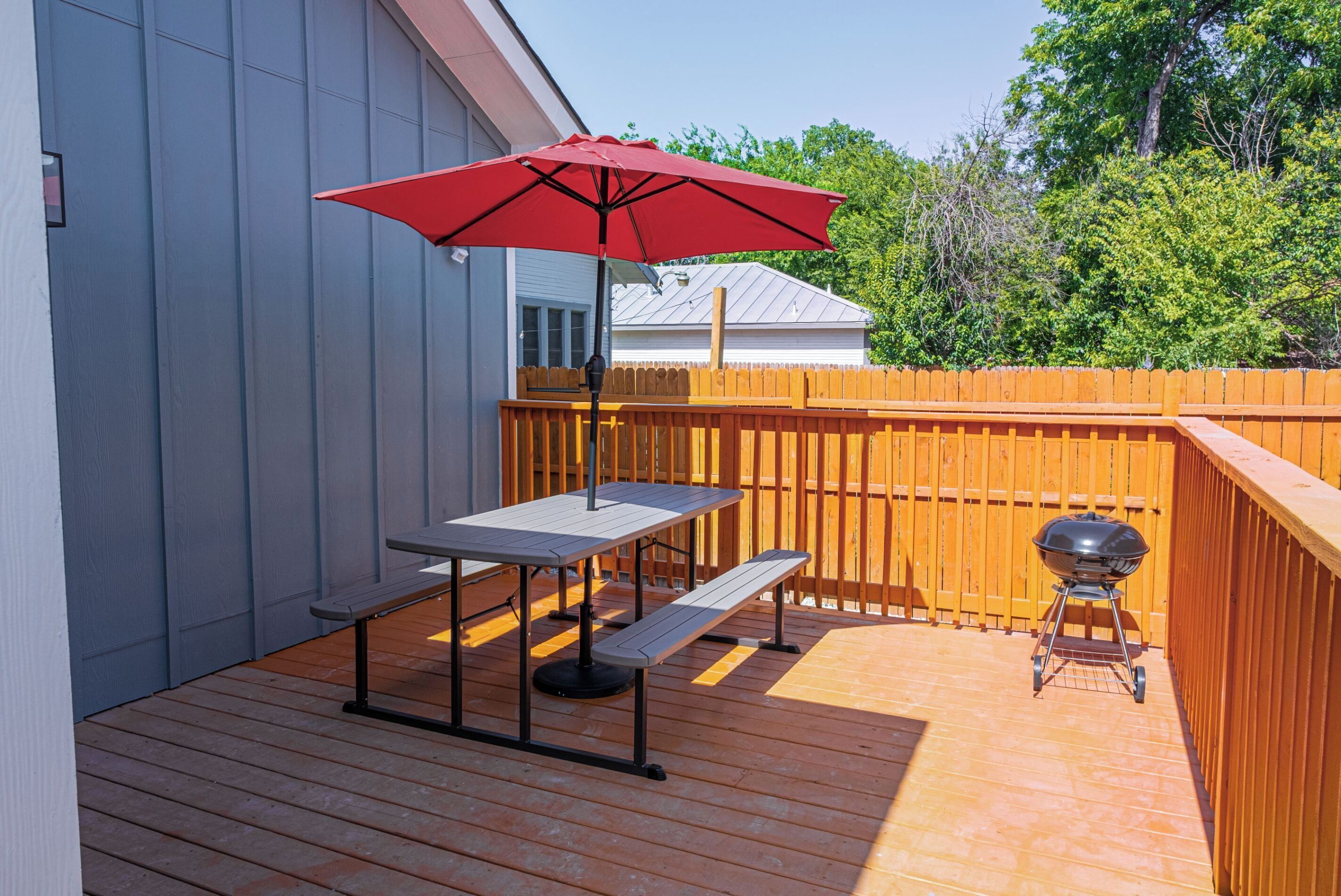 A wooden deck with a table and bench, a red umbrella, and a barbecue grill.