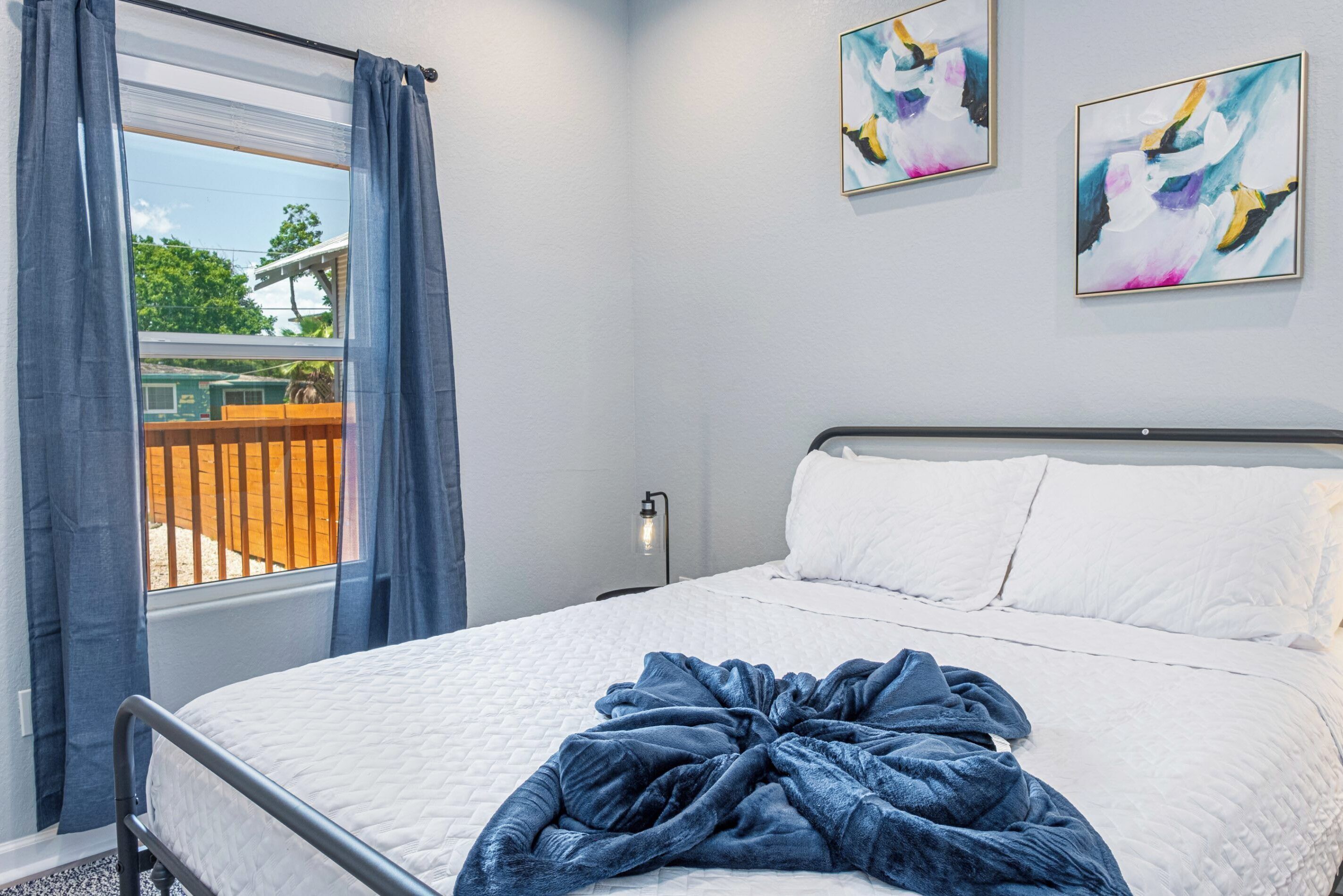 A bedroom with a bed, a window with curtains, and two abstract paintings on the wall.