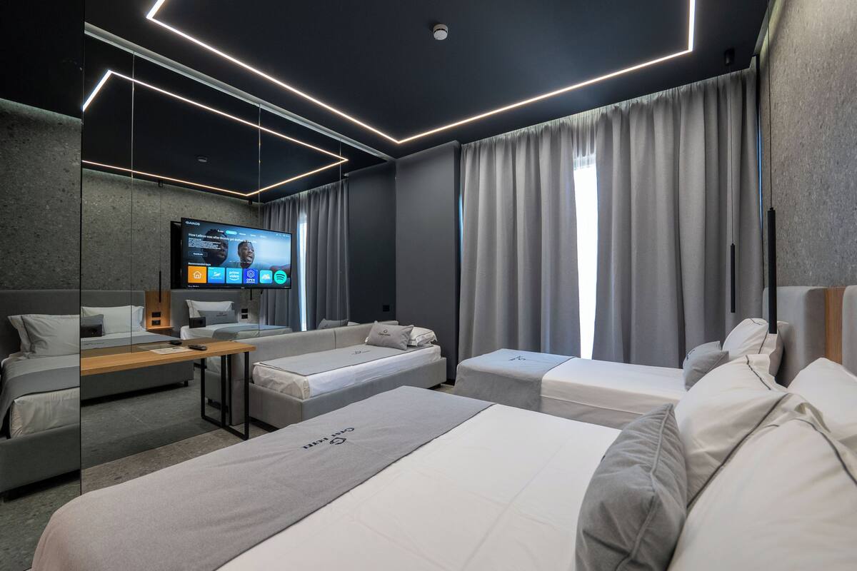 A modern hotel room with a large flat-screen TV, two single beds, a sofa bed, a wooden table, and a large window with curtains.