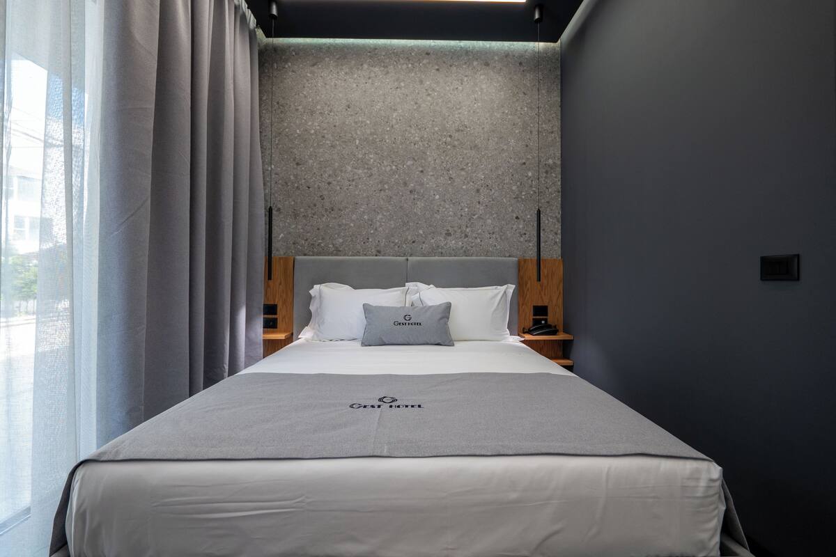 A hotel room with a bed, a grey bedspread, a grey pillow, a wooden headboard, a grey wall, a window with curtains, and a nightstand with a lamp.