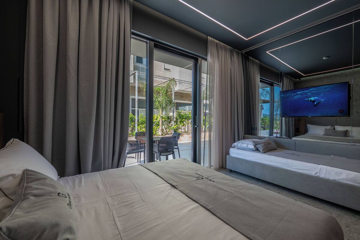 A modern hotel room with a large bed, a sofa, a TV, and a balcony with outdoor seating.