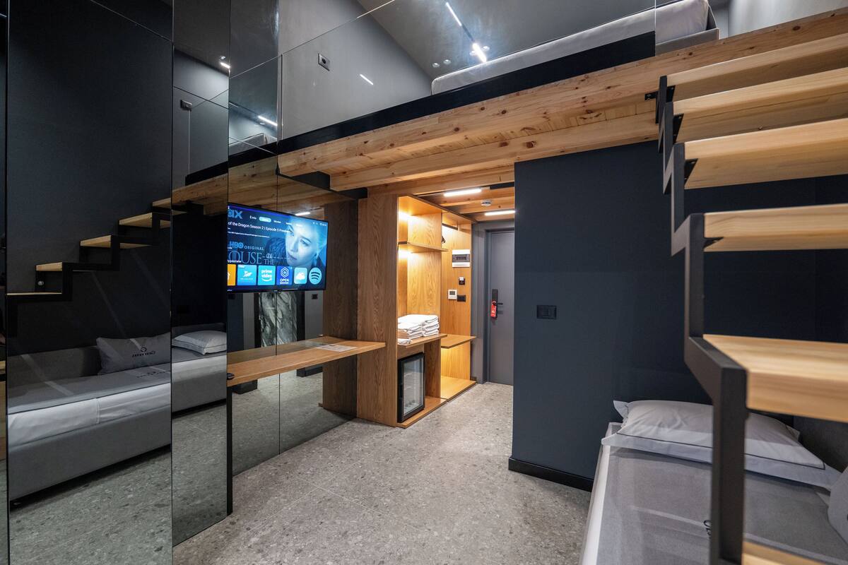 A modern hotel room with a glass partition, a wooden ceiling, a bed, a small seating area, and a television displaying a room service menu.