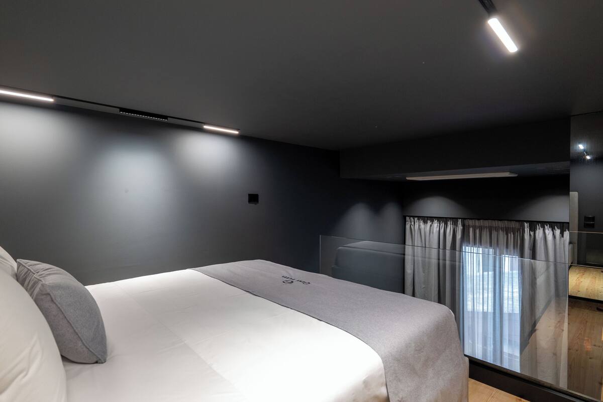 A modern bedroom with a large bed, a bedside table, a mirror, and a window with curtains.