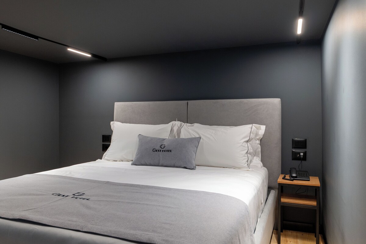 A modern hotel room with a grey headboard, white bedding, a grey pillow with the hotel logo, a bedside table with a phone, and a wall-mounted light.