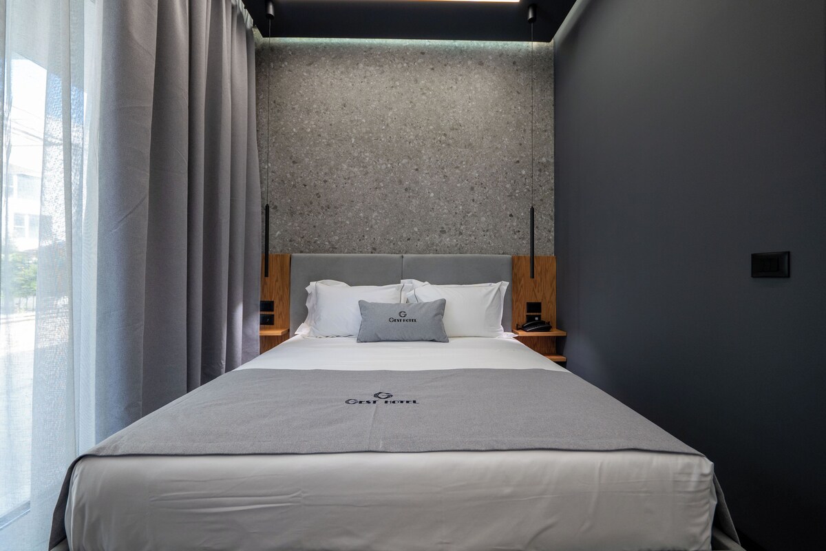 A hotel room with a bed, a grey bedspread, a grey pillow, a wooden headboard, a grey wall, a window with curtains, and a nightstand with a lamp.