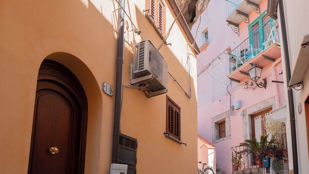 A narrow cobblestone street with buildings on either side, featuring a wooden door and an air conditioning unit on the left.