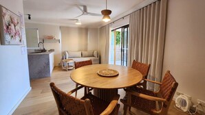 A dining area with a round table and wooden chairs, a ceiling fan, and a view of the outdoors through large windows.