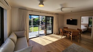 A spacious living area with a sofa, dining table, and chairs, a television, and a sliding glass door leading to a balcony.