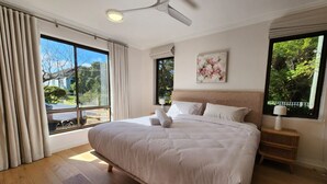 A bedroom with a large bed, a ceiling fan, a window with curtains, and a nightstand with a lamp.