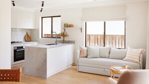 A modern kitchen with a central island, a dining area with a wooden table, and a living room with a sofa and a window.