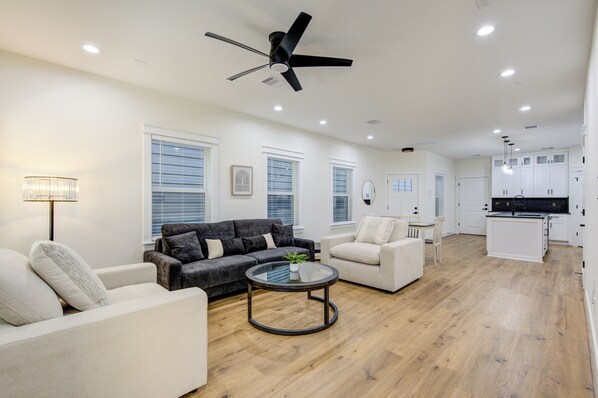 A modern living room with a dark grey sofa, a white sofa, and a glass coffee table. There is a ceiling fan, recessed lighting, and a kitchen area with white cabinets and a black countertop. - Beautiful Luxury Villa near downtown and IAH (Houston)