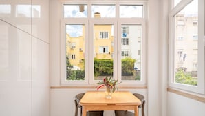 A room with a wooden table and two chairs, a window with a view of a yellow building and greenery, and a white wardrobe. - GuestReady - Bright escape near Santa Apolónia (Lisbon)