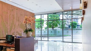 Un área de recepción moderna con un panel de madera, una mesa de recepción, una computadora y un ventanal que ofrece vista a la calle y edificios.
