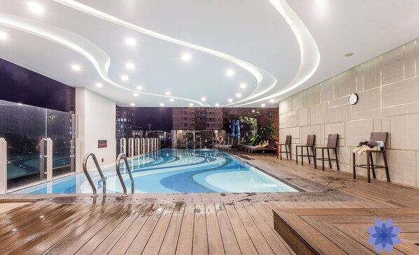 Una piscina interior con terraza de madera, rodeada de sillones de descanso y un diseño de techo moderno.