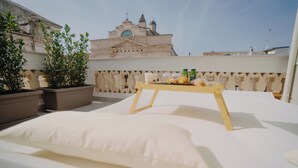 A rooftop terrace with a bed, a small table with food, potted plants, and a building with a clock tower in the background. - Cinquanta Sfumature B&b Luxury Suites (Cassano delle Murge)