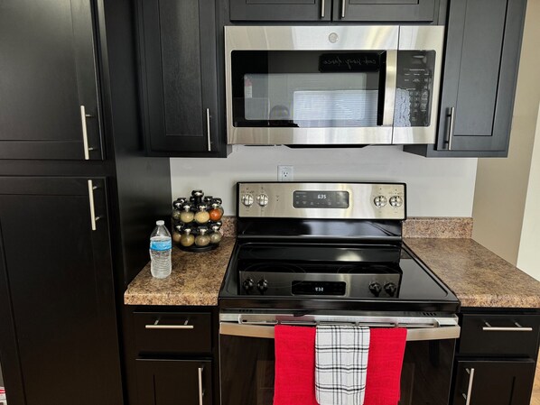 A kitchen with dark cabinets, a stainless steel microwave, a gas stove, and a water bottle on the counter. - Hot Stuff! Pro Cleaned Self Check In Sleeps 10 (Racine)