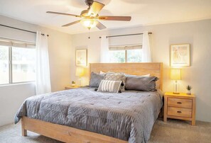 Ein Schlafzimmer mit Bett, Deckenventilator, Nachttischlampen und einem Fenster mit Vorhängen.