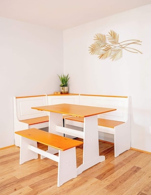 Un banc blanc avec une table et un siège en bois, une petite plante en pot et un élément décoratif mural en feuille d’or.