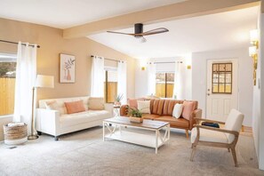 Ein Wohnzimmer mit weißem Sofa, braunem Sofa, Couchtisch, Sessel, Deckenventilator und einem Fenster mit Vorhängen.