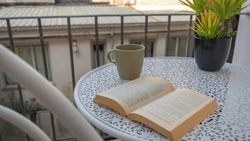 Un livre ouvert sur une table en métal blanc, avec une plante en pot et une tasse verte sur un balcon.