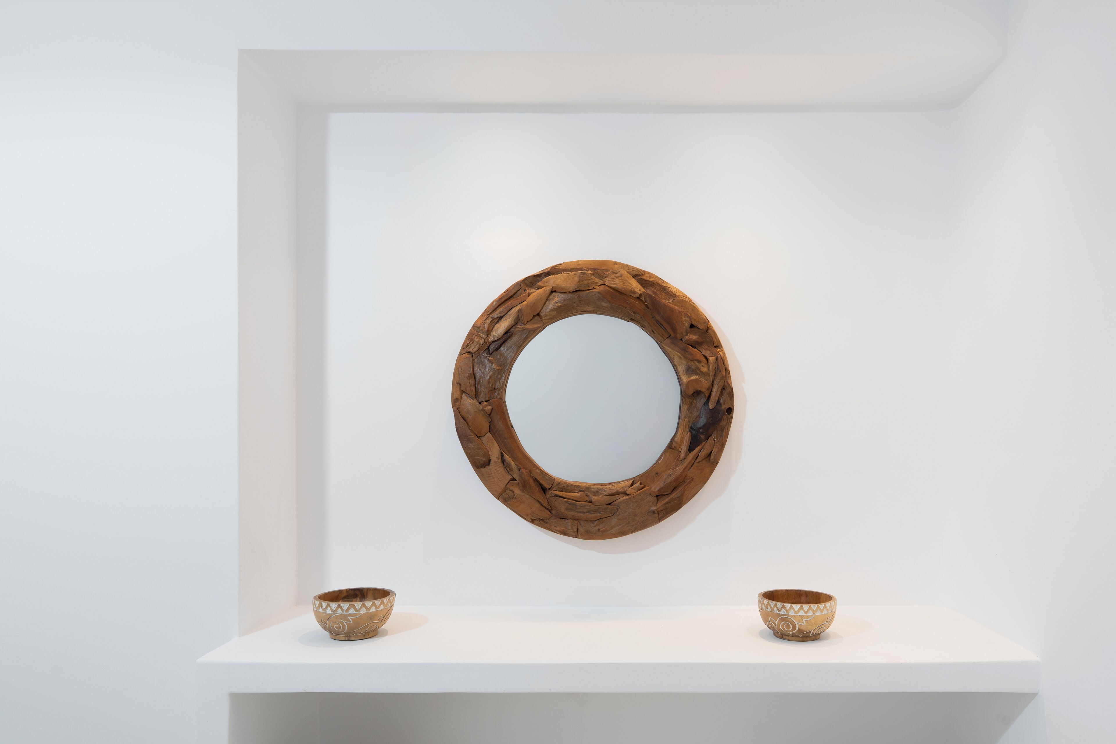 Une pièce au décor minimaliste, avec un miroir rond en bois accroché au mur et deux bols décoratifs posés sur une étagère blanche.