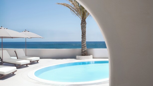 A poolside area with lounge chairs and umbrellas, a palm tree, and a view of the sea. - Casa Frati Suites & Villas (Santorini)