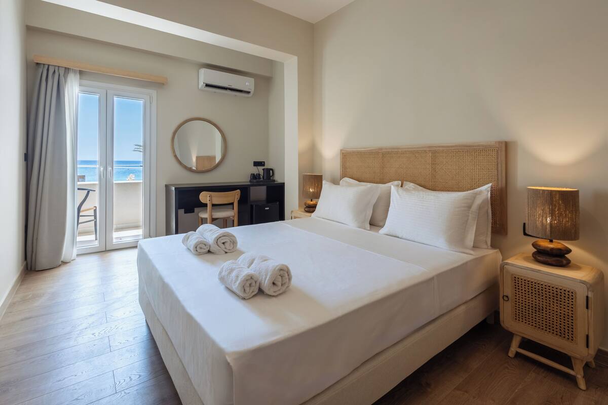 A hotel room with a large bed, a desk with a chair, a round mirror, a bedside table with a lamp, and a view of the sea through a sliding door.